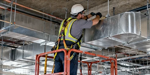 Worker secures suspended HVAC ducts and utility lines beneath a commercial floor slab using ceiling supports.