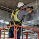 Worker secures suspended HVAC ducts and utility lines beneath a commercial floor slab using ceiling supports.