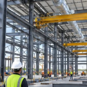 Industrial manufacturing facility under construction with a steel frame, overhead crane rails, and a crew installing building systems.