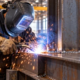 Certified welder joins structural steel in an industrial shop while an inspector checks the completed weld.