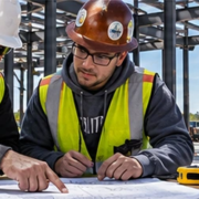 Foreperson shows an apprentice how to measure a commercial building layout against a plan, demonstrating hands-on construction training.