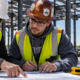 Foreperson shows an apprentice how to measure a commercial building layout against a plan, demonstrating hands-on construction training.