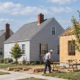 A builder frames a mid-century house while modest detached homes line a suburban street, reflecting postwar neighborhood construction.
