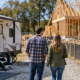 Homeowners stay in a travel trailer outside a fenced house build while their new home is framed nearby.