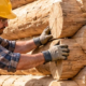 Craftsperson fits rounded interlocking logs at the corner of a small cabin on a stone foundation while building traditional log walls.