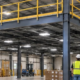 Industrial mezzanine platform adds an intermediate level above the warehouse floor, with stairs, beams and guardrails visible.