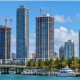 Miami-Dade waterfront skyline with active residential and commercial towers, construction cranes, and palm trees in tropical daylight.