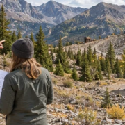 Land surveyor and owner review a site plan beside a proposed cabin on rugged former mining land with boundary markers.
