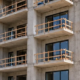 Multi-family concrete apartment building under construction with repeated balcony bays, perimeter scaffolding, and workers on the structure.
