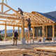 Builders install timber posts and roof trusses on a pole barn house frame in a rural setting.