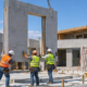 Crane lifts a precast concrete wall panel onto the foundation of a modern single-family house as the crew guides it into place.