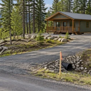 Wooded recreational parcel with a small cabin on a cleared pad, gravel access road, survey markers, and a visible drainage swale.