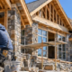 Mason fits natural stone pieces onto a house exterior as the stone walls rise beneath the timber roof framing.
