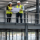 Architect and site manager review plans on a steel mezzanine overlooking a warehouse floor, showing an intermediate level that uses the building’s vertical space.