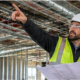 Systems engineer points to overhead building services while an architect reviews plans at an active construction site.