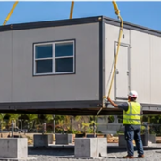 Crane lowers a prefabricated classroom module beside a school while workers guide the installation.
