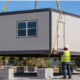 Crane lowers a prefabricated classroom module beside a school while workers guide the installation.