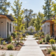 Builder finishes a compact modular home beside a landscaped shared walkway in a small transitional housing community.