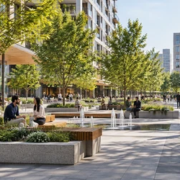 Pedestrian plaza with shaded seating and greenery links contemporary buildings in an active urban district.
