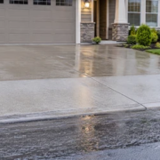 Concrete driveway apron between a residential garage and street, directing runoff toward a curbside drain