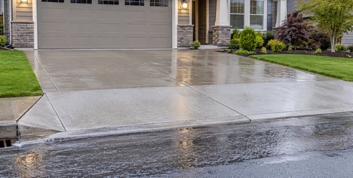 Concrete driveway apron between a residential garage and street, directing runoff toward a curbside drain
