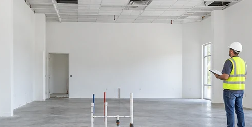 Empty commercial white-box suite with finished white walls, a basic ceiling grid, concrete floor, utility stubs, and a contractor inspecting it for tenant fit-out.