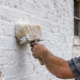 Craftsperson brushes limewash over a textured masonry wall, with the transition from bare brick to white finish visible.